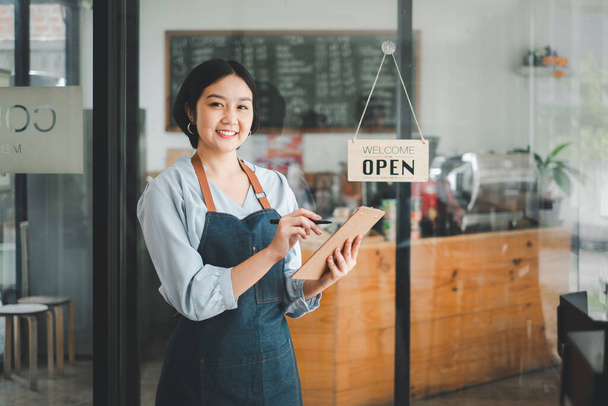 Beautiful young barista woman in apron holding order paper and standing in front of the door of cafe with open sign board. Business owner startup concept. - 写真・画像
