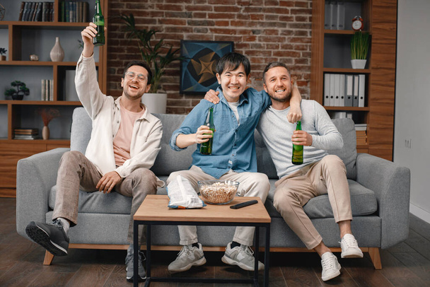 Front view of male friends gathered at home to watch a football match. Men sitting on the couch in front of the big screen TV and eating snacks. Men actively support their favorite team and feeling - Foto, immagini
