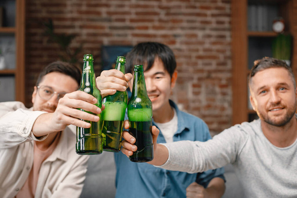 Focus on a bottles of beer. Front view of male friends gathered at home to watch a football match. Men actively support their favorite team and comment on the match - Zdjęcie, obraz