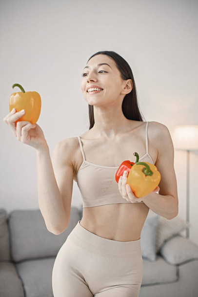 Slim and beautiful woman wearing beige leggins and top and holding fresh red and yellow paprika in hands. Young girl with fit body posing at home. Sport, fitness, diet, nutrition and health care - Foto, imagen
