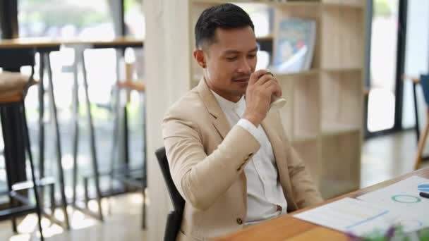 Portrait of an Asian business man drinking coffee while working with a computer and financial statements documents on his desk. High quality 4k footage - Footage, Video