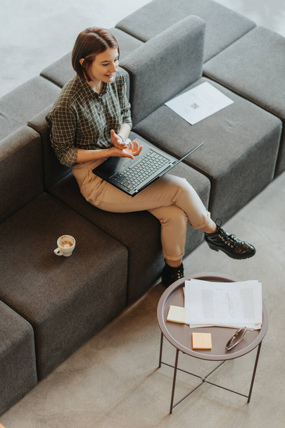 Kurzhaarige Business-Girl arbeitet auf ihrem Laptop, während sie auf einem grauen Sofa mit einer Tasse Kaffee neben sich sitzt. Sie unterhält sich mit ihren Kollegen und lächelt - Foto, Bild