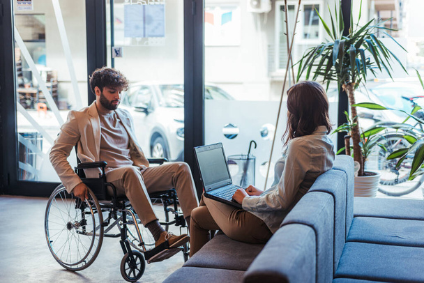 Brunette female employee discussing with her handicapped male colleague in wheelchair while working on lap top - Photo, Image