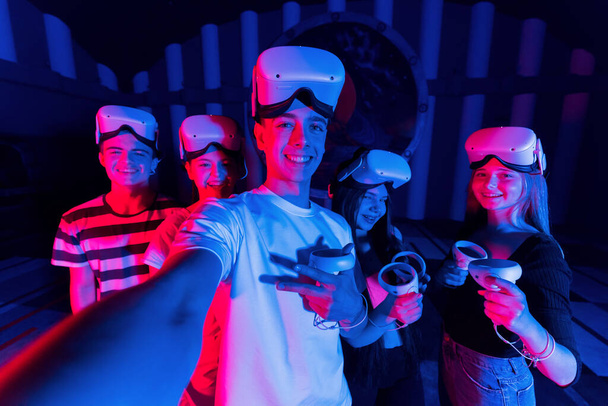 Uma equipe de jovens amigos tirando selfie ao usar equipamentos de realidade virtual em uma arena. Luzes de néon - Foto, Imagem