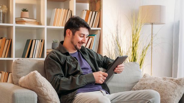 Gadget leisure. Internet entertainment. Watching content. Joyful man browsing digital tablet online relaxing on cozy couch in living room at home. - Foto, Imagen