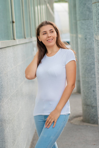 A young woman in a white T-shirt poses against the backdrop of a wall and columns. Mock-up. - Фото, изображение