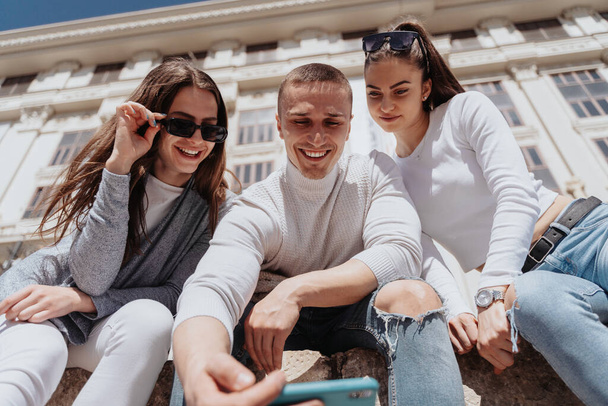 Acercamiento de tres amigos tomando una selfie mientras están sentados afuera frente al edificio blanco - Foto, imagen