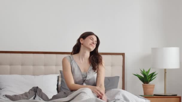 Μια νεαρή γυναίκα σηκώνεται από το κρεβάτι και τεντώνει τους ώμους της μετά από έναν μακρύ ύπνο. Ξυπνώντας μια νεαρή μελαχρινή θαυμάζει το φως του ήλιου από το παράθυρο, ενώ κάθεται στο κρεβάτι. Υψηλής ποιότητας υλικό FullHD - Πλάνα, βίντεο