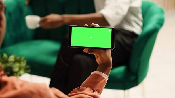 Afroamerikanerin mit Chroma-Schlüssel, grünem Bildschirm, Smartphone-Attrappe im Querformat auf gemütlichem Sofa sitzend. Touristen warten in eleganter Hotel-Lounge auf Einchecken - Foto, Bild