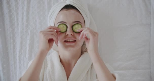 Vtipná šťastná mladá žena v županu s ručníkem na hlavě zakrývající oči okurkami propagující svěží zdravou koncepci péče o pleť. žena držící organickou lázeňskou masku smějící se na kameru - Záběry, video