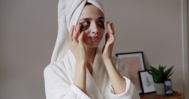 Νεαρή όμορφη γυναίκα σε ένα μπουρνούζι και μια πετσέτα στο κεφάλι με υγιές δέρμα γελάει χαρούμενα εφαρμόζει καφέ κηλίδες για να μειώσει σακούλες ματιών. Αναζωογόνηση του δέρματος περιποίηση προσώπου γυναίκα πρόσωπο. - Πλάνα, βίντεο