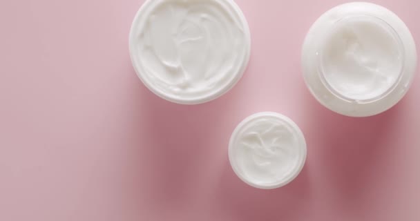 Draufsicht Weiße Flaschen mit Hautpflegecreme, für Kosmetik auf rosa Hintergrund im Studio drehen. Kosmetiköl auf Basis natürlicher Inhaltsstoffe, Peeling, Tonic, Körperpflege. Gruppe verschiedener Kosmetikflaschen - Filmmaterial, Video