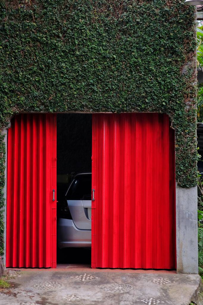 赤いローリングドアとツタの葉に覆われた壁を持つ車のガレージ. - 写真・画像