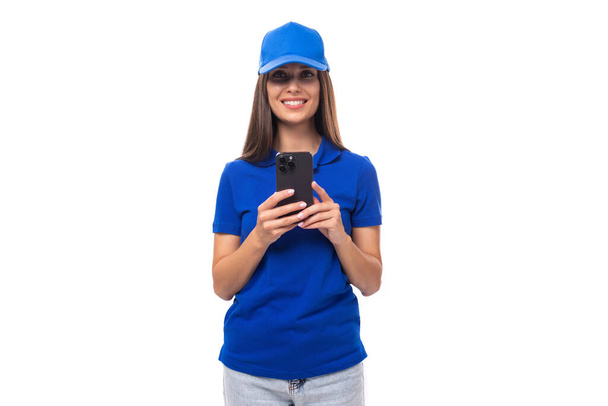 vrij vriendelijke jonge promotor vrouw in blauw katoen t-shirt en pet met smartphone. - Foto, afbeelding