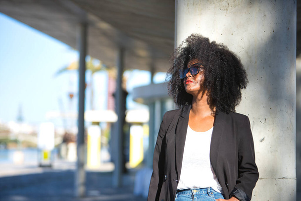 Junge, schöne, schwarze Frau mit Afrohaaren, Jacke und Sonnenbrille, die an eine Betonsäule gelehnt ist und die Sonnenstrahlen empfängt. Konzept Empowerment, Erfolg, aktuell, modern. - Foto, Bild