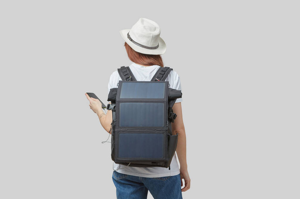 Girl carrying backpack with flexible solar panel attached to smartphone in her hands, isolated on light gray background. Travel and alternative energy concept. - Photo, image