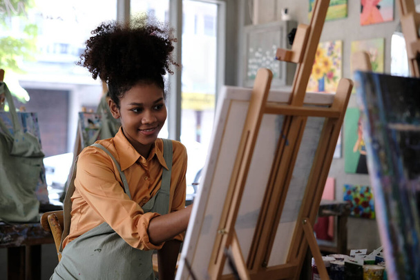 Kreatywna młoda Afroamerykanka nosząca fartuch obraz z pędzlem na płótnie w pracowni sztuki. - Zdjęcie, obraz