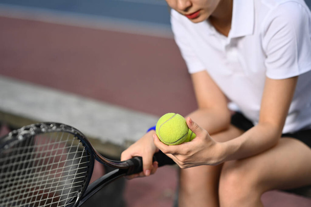 Όμορφη νεαρή αθλήτρια που κρατάει ρακέτα καθισμένη στον πάγκο στο γήπεδο τένις. - Φωτογραφία, εικόνα