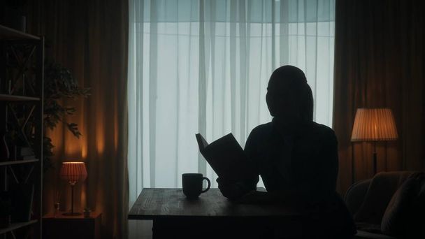 A woman reads a book sitting at a table with a cup of coffee, tea in the room on the background of the window. Dark silhouette of a woman with an open book in her hand - Фото, зображення