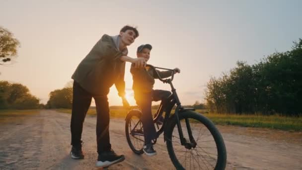 Irmãos alegres aprendem a andar de bicicleta ao pôr-do-sol. O irmão mais velho ensina o mais novo. Feliz conceito de família. Trabalho de equipa. As crianças sonham. Imagens 4k de alta qualidade - Filmagem, Vídeo