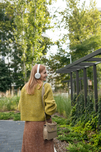 Back view of young blonde Caucasian woman listening the music while walking through the city. High quality photo - Фото, изображение