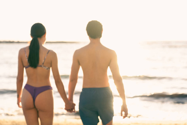 Handsome man wearing swimsuit with beautiful woman walks on the beach in a sunny day, holding hands, during holidays. Young adults caucasian couple travel together a romantic vacation relaxing outdoor - Photo, Image