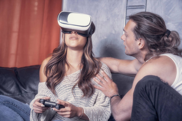 Cheerful modern couple of caucasian guys play together in living room of their studio house. Male guy enjoyng life with a girl playng videogames with cell phone goggles holding joypad having fun - Photo, Image
