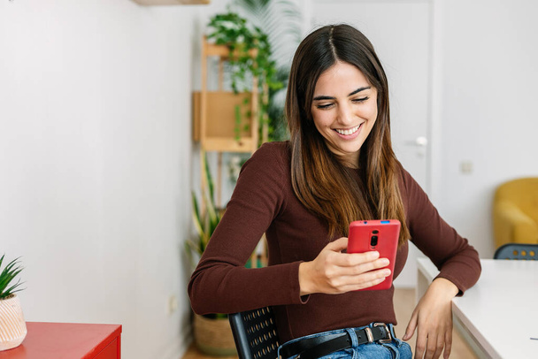 Szczęśliwa kobieta uśmiechnięta podczas korzystania z telefonu komórkowego relaks w domu. Millennial student sprawdzanie aplikacji social media na smartfonie podczas nauki online na laptopie. - Zdjęcie, obraz