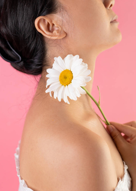 Κοντινό πλάνο της χαρούμενης όμορφης νεαρής Ασιάτισσας που ποζάρει σε ροζ φόντο, αγγίζει το λαιμό με λευκό λουλούδι μαργαρίτα και χαμογελά. - Φωτογραφία, εικόνα