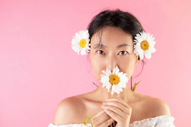 Primer plano de feliz alegre bonita mujer asiática posando sobre fondo rosa, tocando la cara con flor de margarita blanca y sonriendo. - Foto, Imagen