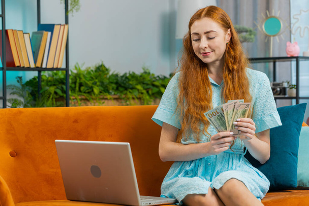 Suunnittelubudjetti. Rikas onnellinen punapää nainen laskee rahaa käteisellä käyttää kannettavaa tietokonetta laskea kotimaan laskuja kotona. Iloinen tyttö tyytyväinen tuloihin, säästää rahaa suunniteltuun lomaan, lahjoihin - Valokuva, kuva