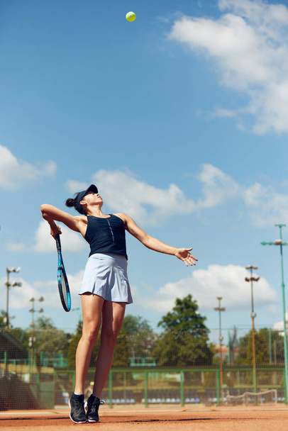 Full-length dynamic image of young athletic woman, tennis player in uniform during game, playing at open air stadium, court on warm day. Concept of sport, hobby, active lifestyle, health, strength, ad - Foto, imagen