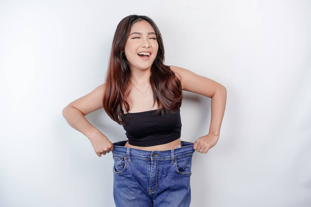 Diet and weight loss concept. A young Asian woman in oversized jeans isolated on a white background. - Foto, imagen