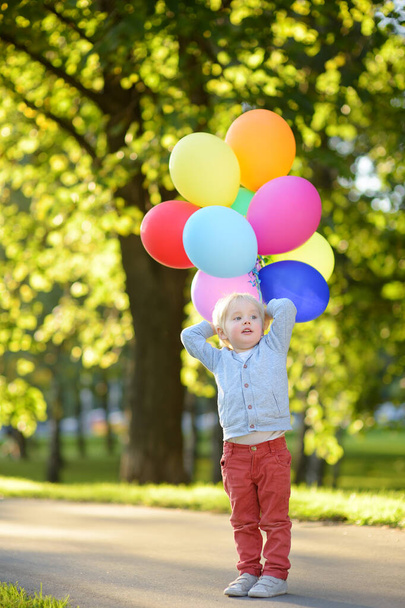 Little boy having fun on celebrating birthday party. Happy child with bunch of bright multicolor balloons. Preschoolers or toddlers baby birthday party in sunny park. Summer outdoor festival for kids - Photo, Image