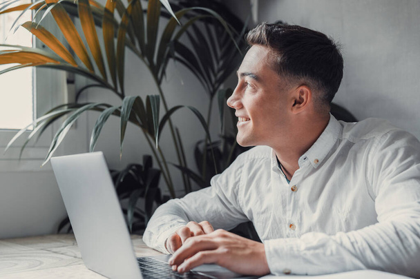 przystojny młody biznesmen z laptopem przy biurku, mężczyzna patrząc i myśląc z uśmiechem  - Zdjęcie, obraz