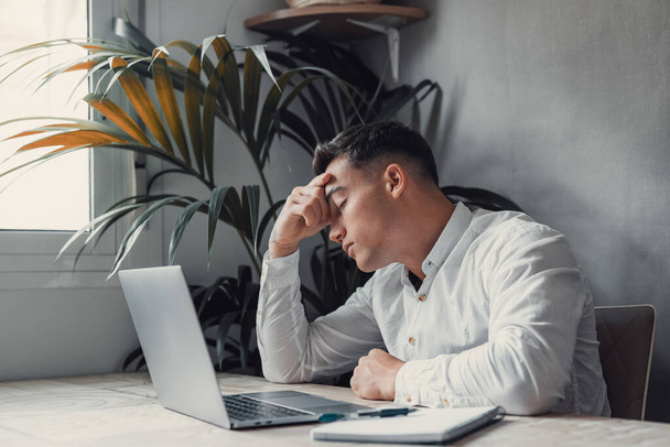 Εξαντλημένος και κουρασμένος άνθρωπος στο γραφείο με φορητό υπολογιστή. επιχειρηματίας με κλειστά μάτια και χέρι στο κεφάλι  - Φωτογραφία, εικόνα
