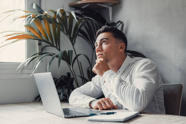 joven hombre de negocios guapo con el ordenador portátil en el escritorio, hombre mirando hacia arriba y pensando con la mano en la barbilla  - Foto, imagen