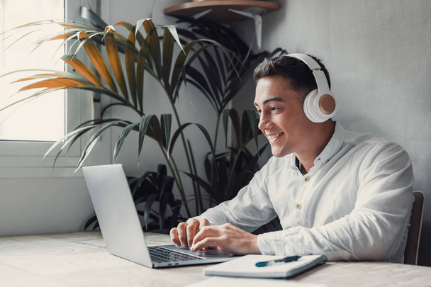 beau jeune homme d'affaires avec ordinateur portable au bureau, portant des écouteurs et souriant  - Photo, image
