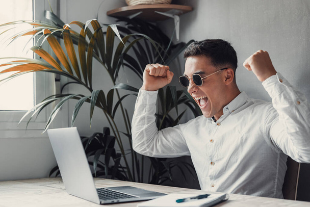 Muž ve slunečních brýlích u stolu, dívá se na laptop a oslavuje. úspěšné podnikatelské gesto s rukama  - Fotografie, Obrázek