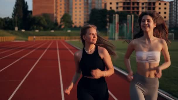 Twee mooie meisjes joggen bij zonsondergang in het stadion. Jonge blanke vrouwen die samenwerken, glimlachend. Een gevoel van vrijheid tijdens het rennen. Cardio-concept. - Video