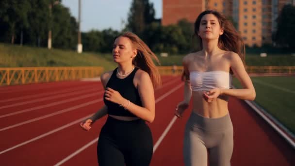 Nuori urheilullinen hoikka nainen juoksee stadionilla, kaksi naispuolista ystävää kuntoilee ulkona auringonlaskun aikaan kesällä - Materiaali, video