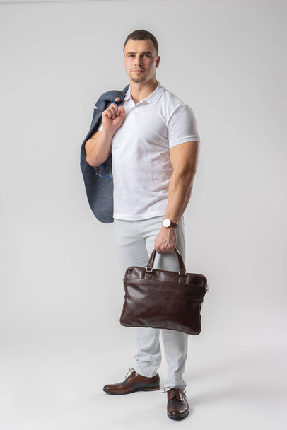 Un hombre de complexión atlética con ropa ligera casual sostiene un maletín de cuero y una chaqueta en sus manos. Modelo posando sobre un fondo blanco. - Foto, imagen
