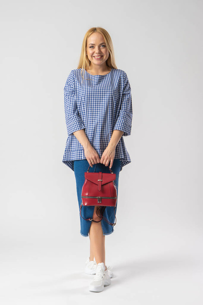 A young blonde woman in a casual denim skirt and a plaid blouse stands and holds a red backpack bag in her hands, looking at the camera. Model posing on white background - Foto, immagini