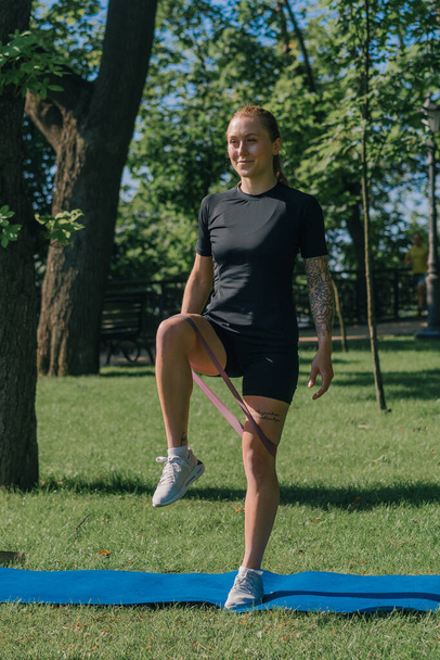 mladá sportovkyně dělá fyzická cvičení na sportovní matrace trénink s gumičkou na nohou venku v parku zdravý životní styl koncept - Fotografie, Obrázek