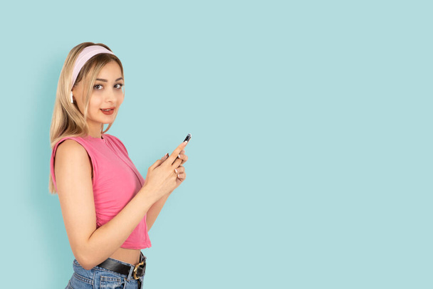 Using smartphone, side view portrait of young attractive girl using smartphone. Posing isolated light blue background, copy space. Using mobile phone, application, messaging. Headset device accessory. - Photo, Image