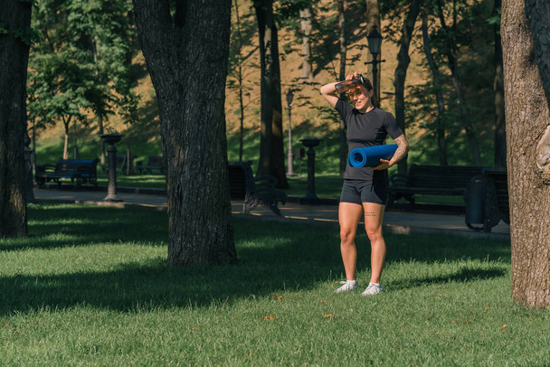 młoda sportowiec dziewczyna trzyma karemat i butelkę wody w rękach przed treningiem na zewnątrz w parku kładzie mat na trawie zdrowy styl życia koncepcja - Zdjęcie, obraz