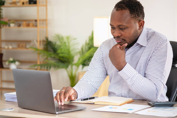 Afroamerykański biznesmen czarnego pochodzenia siedzi uśmiechnięty i robi raporty finansowe i studiuje coroczną analizę zysków Księgowy sprawdzający status finansowy firmy jest w biurze. - Zdjęcie, obraz