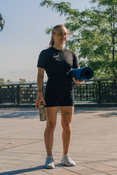 портрет молодой улыбающейся спортсменки с гирей и бутылкой воды в руках перед началом тренировки на открытом воздухе в парке - Фото, изображение