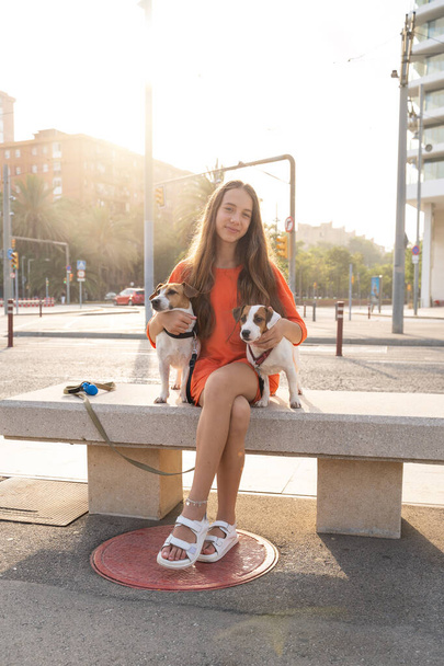 Adorable teenager girl sitting on the bench with her two Jack Russell terriers dogs. Bright orange color casual sporty clothes. Summer evening time walking with dogs in big city.  - Fotografie, Obrázek