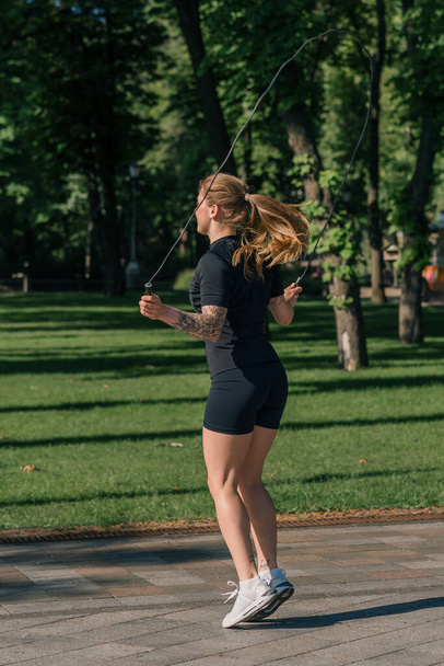 Fitness young sportsman girl with tattoos in the morning doing sports jumping rope during exercise outdoors in the city in health park - Foto, Imagen